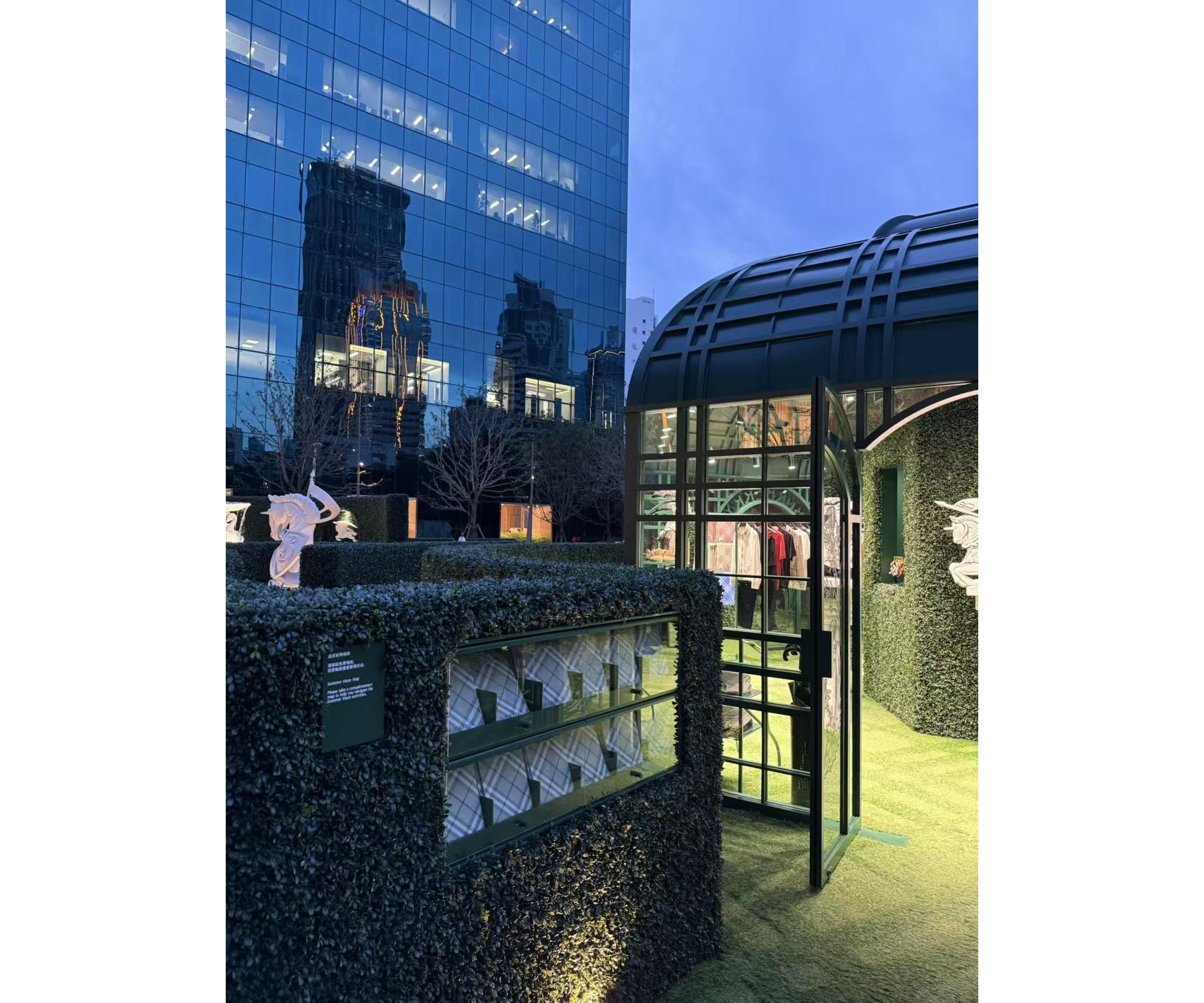 黄昏时分的 Burberry 快闪店外观,四周设有绿篱墙和玻璃展示柜。